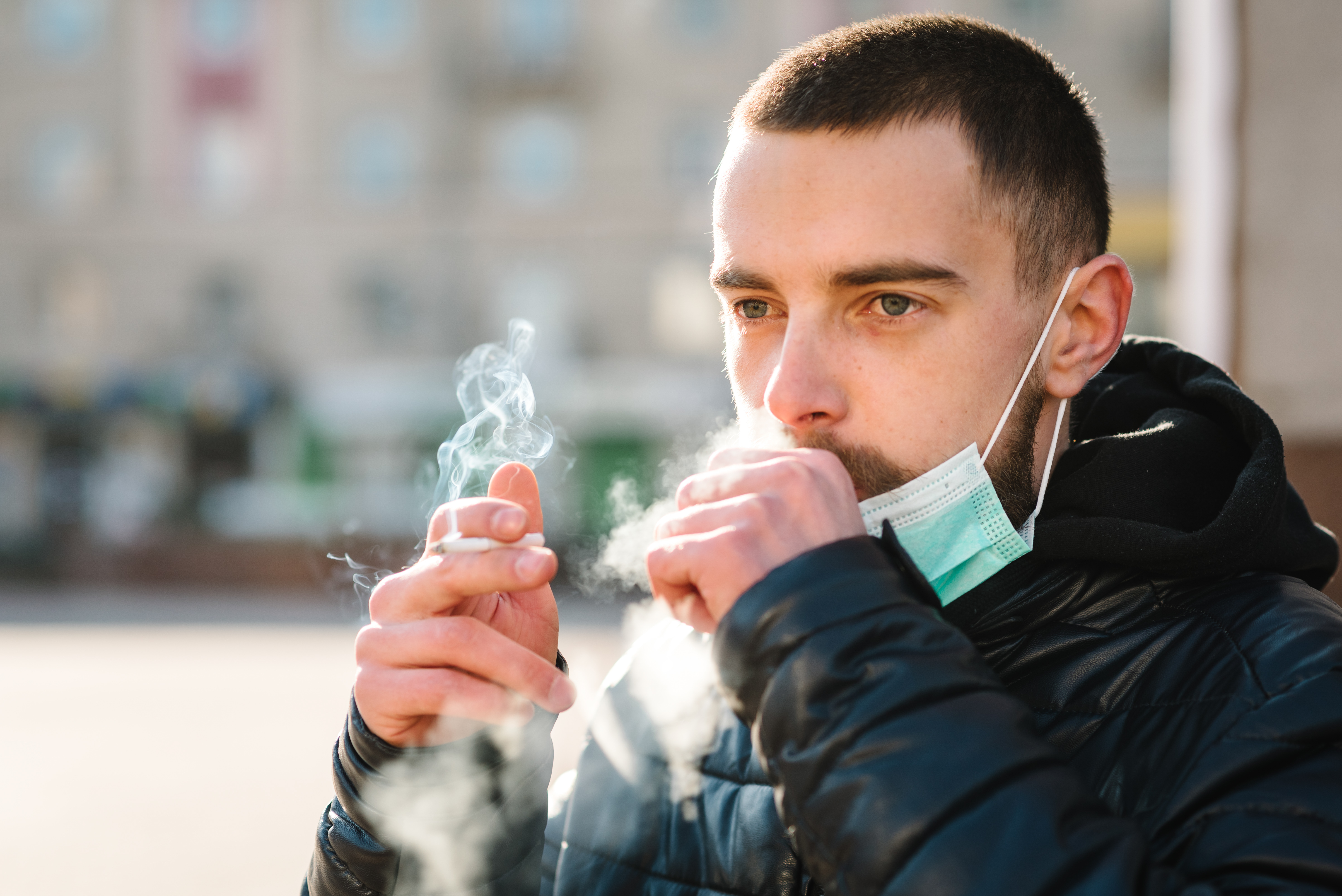Closeup man with mask during COVID-19 pandemic coughing and smoking a cigarette at the street